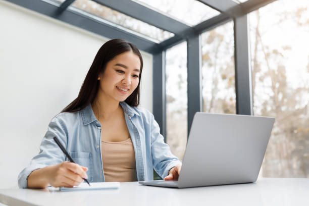 Girl student looking at the laptop and preparing an essay in a notebook